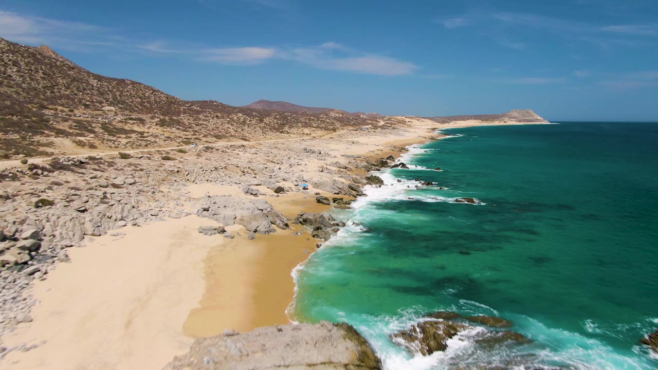 Wide aerial frame at Buzzards Beach showing more open sand in the lower left, rocky shoreline through the center, deep blue ocean to the right, and no visible people.