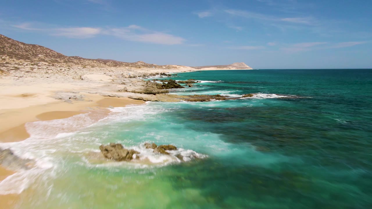Aerial view of turquoise waves breaking beside sandy beach and rocks.