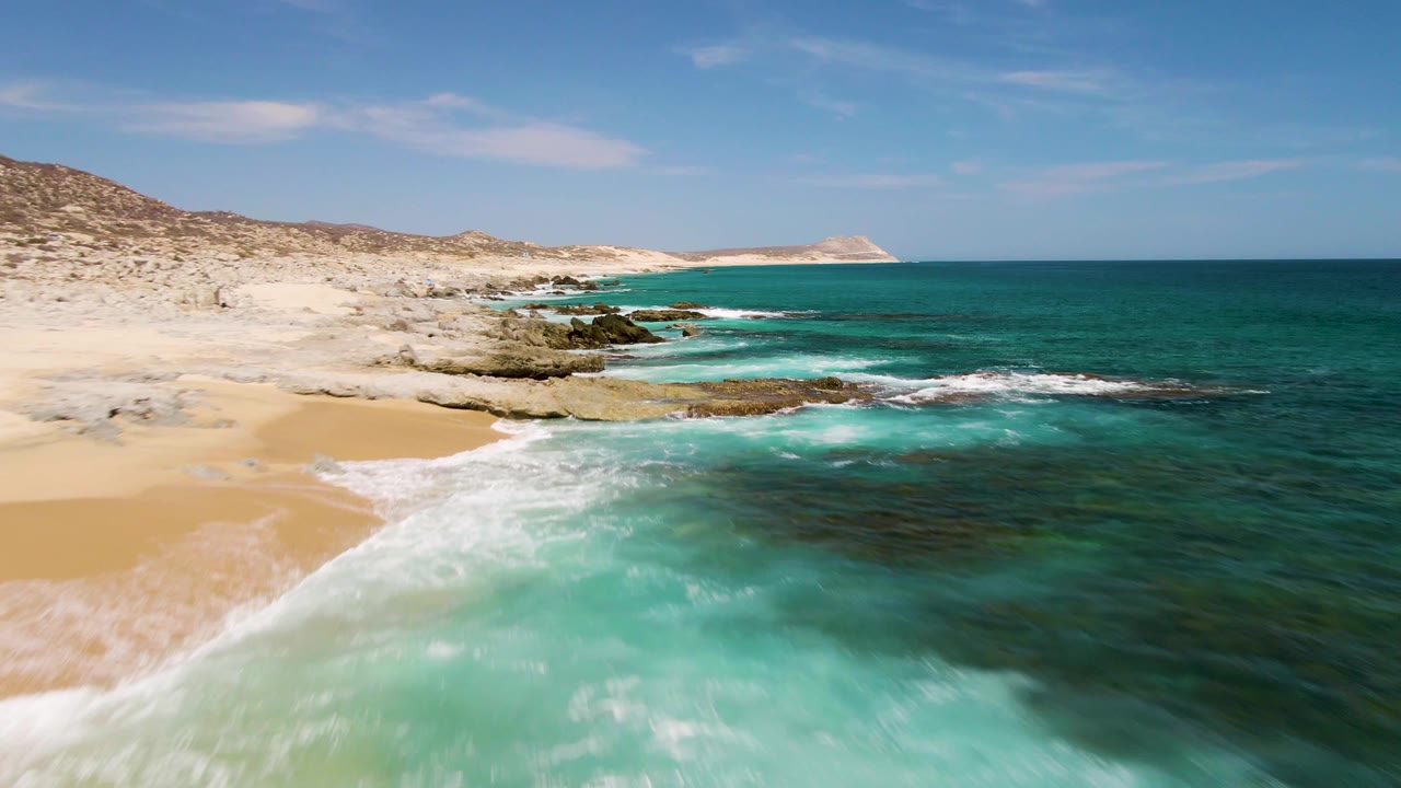 Aerial frame from Buzzards Beach, Baja California Sur, with a sandy cove on the left, clear turquoise water in the foreground, rocky shelves through the center, and no visible people.
