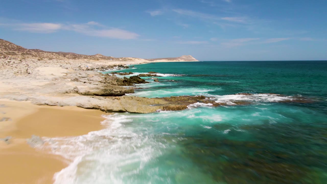 At Buzzards Beach, Baja California Sur, white surf pushes across the sand near exposed rocks while darker reef patches show beneath the green-blue water, with no visible people.
