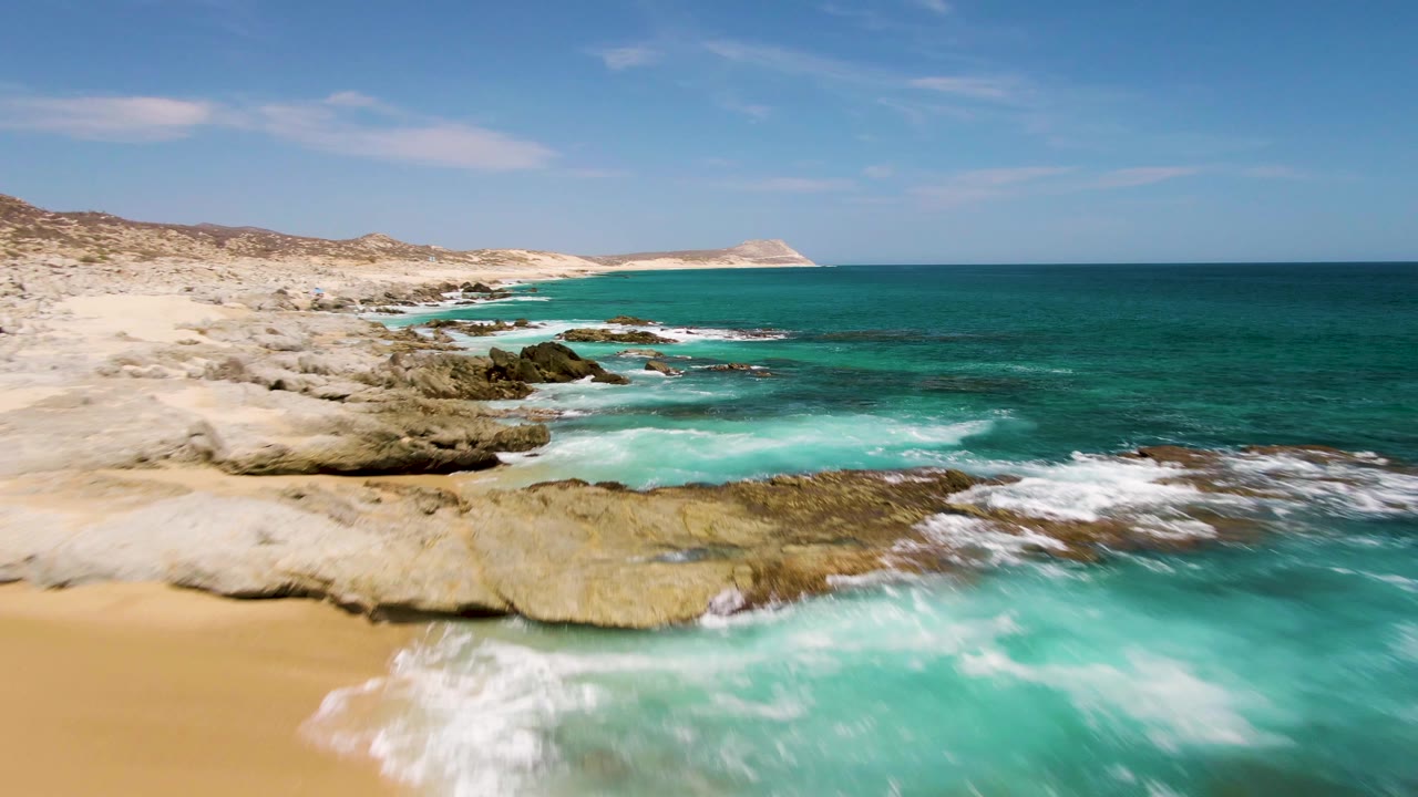 Aerial view along Buzzards Beach, Baja California Sur, where waves break over a rocky shelf beside tan sand, with the shoreline stretching toward a distant point and no visible people.