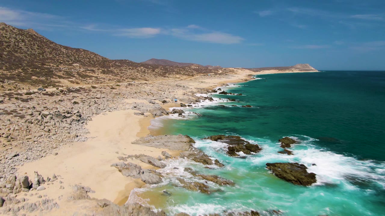 Aerial frame at Buzzards Beach with white waves spreading farther around the foreground rocks, pale sand on the left, dry rocky hills beyond, deep blue water to the right, and no visible people.