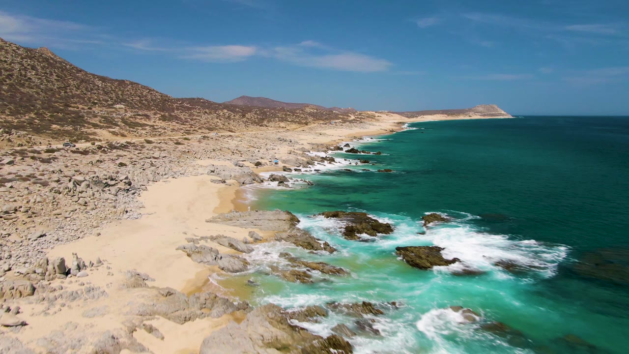 Aerial view of Buzzards Beach showing a broader sweep of white foam across the rocky shallows, tan sand along the left cove, arid hillside behind it, open blue ocean, and no visible people.