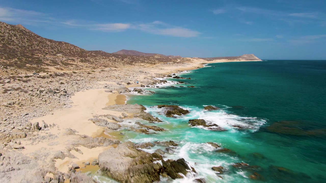 Aerial frame over Buzzards Beach with the sandy beach more open on the left, surf lines wrapping the central rocks, darker blue water beyond the shallows, dry hillside inland, and no visible people.