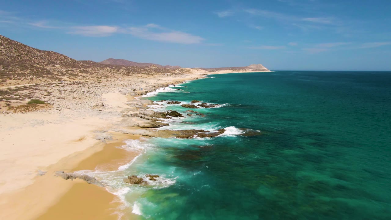 Aerial frame over Buzzards Beach, Baja California Sur, showing pale sand, turquoise water, dark rocks in the surf, low hills, and no visible people.