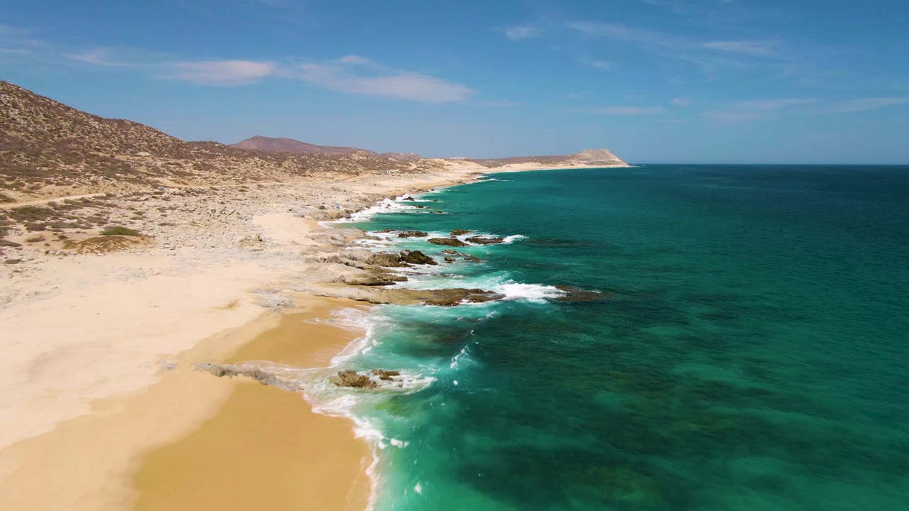 Aerial view at Buzzards Beach with the surf line slightly farther along the sand, green-blue shallows, dark rocks, distant low hills, and no visible people.