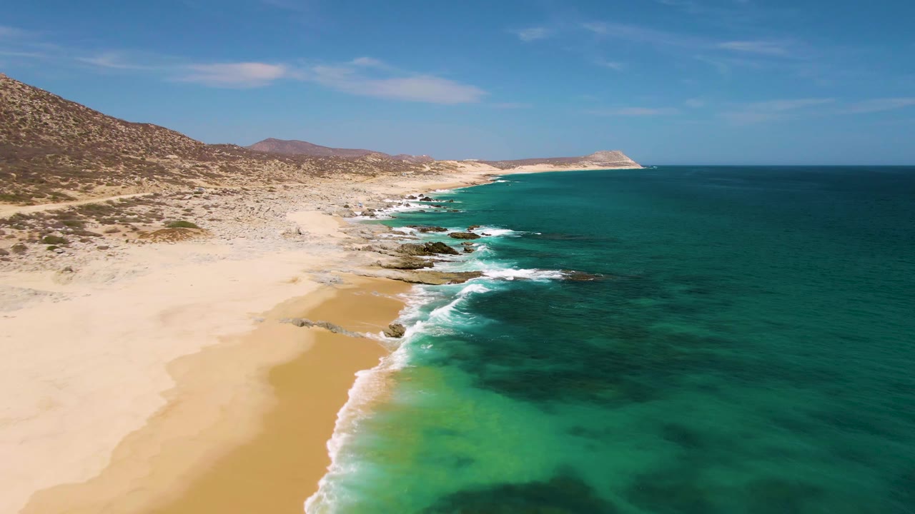 Wide aerial coastal frame with sandy beach on the left and dark blue ocean on the right.