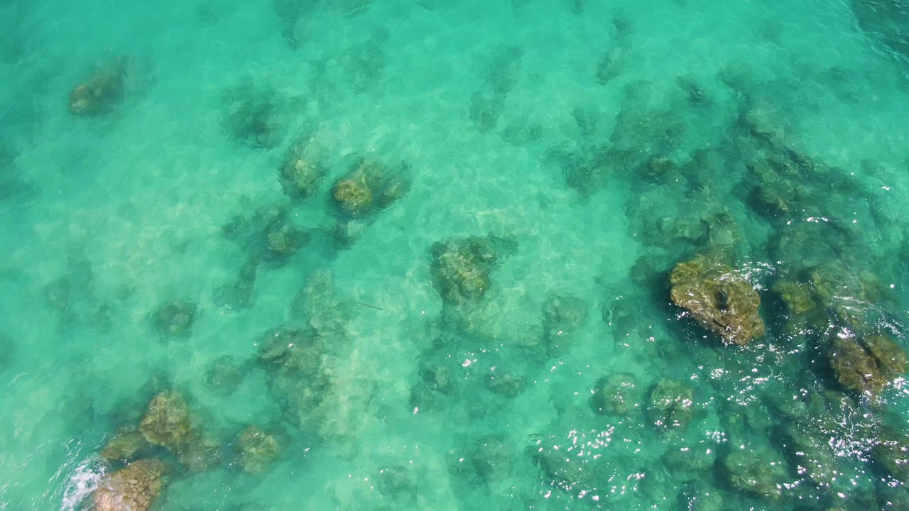 Clear turquoise ocean at Buzzards Beach shows darker underwater rock and coral patches through soft ripples, with no visible people.