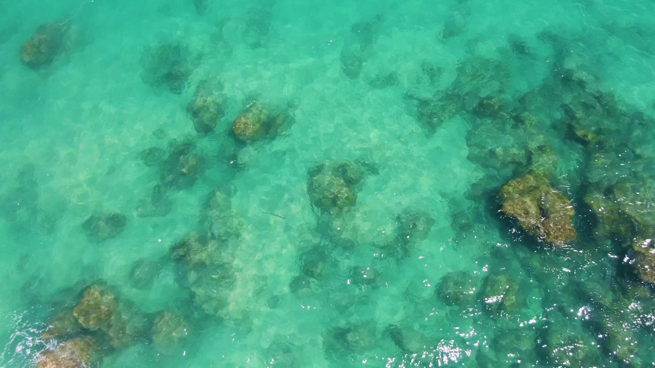 Overhead turquoise water at Buzzards Beach reveals larger submerged rock clusters near the lower frame edge, light ripples, and no visible people.