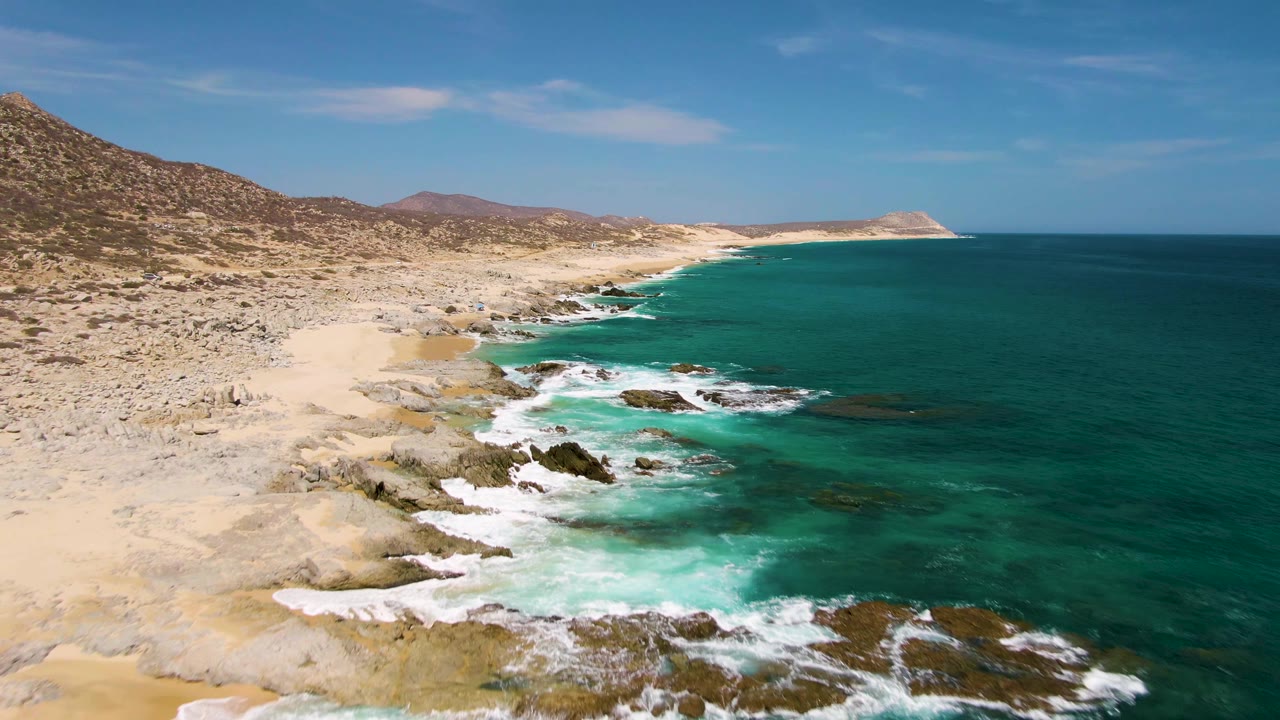Aerial view at Buzzards Beach with white water pulling around exposed rocks, a sandy cove on the left, open teal ocean on the right, and no visible people.