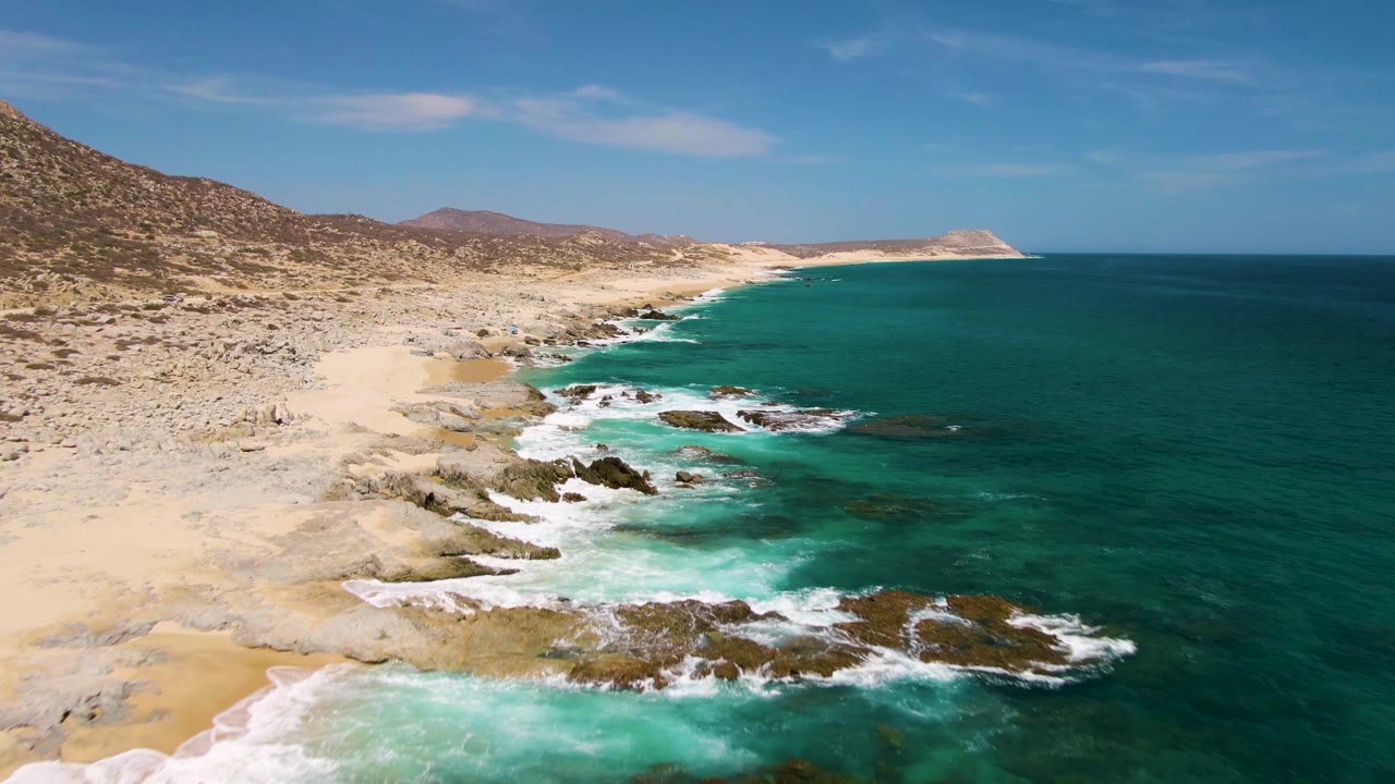 Aerial coastal frame from Buzzards Beach showing a wider stretch of arid shore, foamy surf over dark rocks, deep green-blue water, and no visible people.