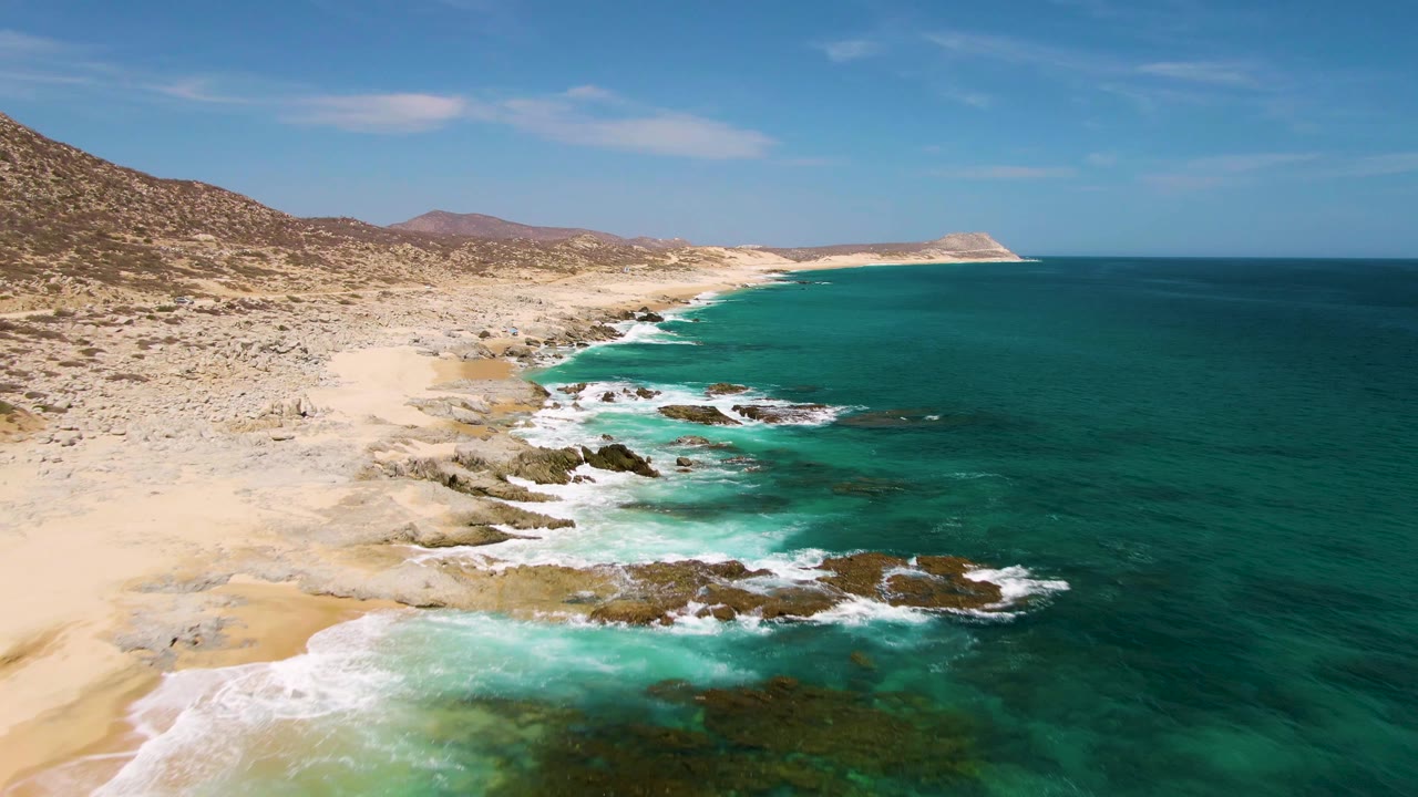 Aerial frame over Buzzards Beach with the sandy shore curving through the left side, rocky outcrops in the shallows, broad teal water to the right, and no visible people.