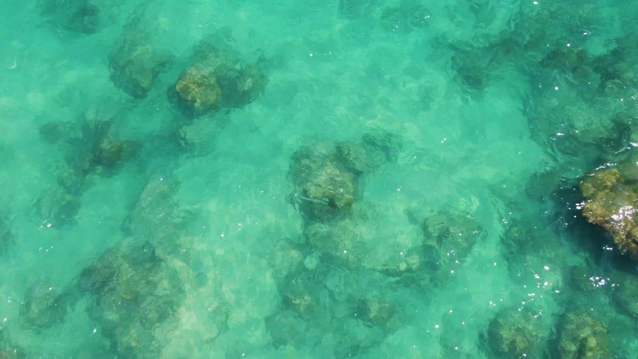 A top-down Buzzards Beach frame with larger brown rocks visible below turquoise water and small sun reflections near the surface, no visible people.