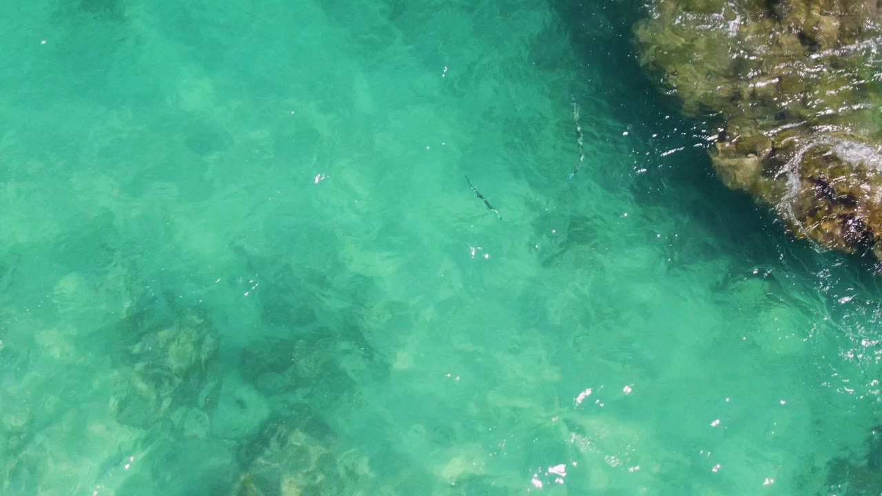 Clear turquoise water at Buzzards Beach shows darker submerged rock patches across the center and a rough reef edge in the upper right, with no visible people.