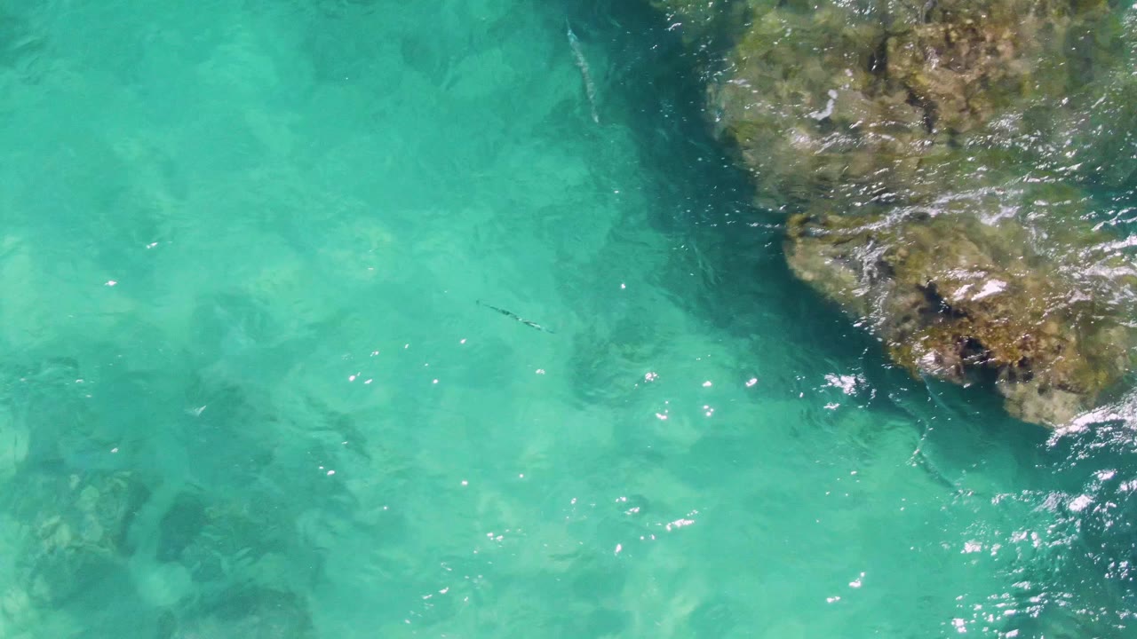 Buzzards Beach shallow water appears bright turquoise below a larger section of rocky outcrop on the right, with faint underwater rocks and no visible people.