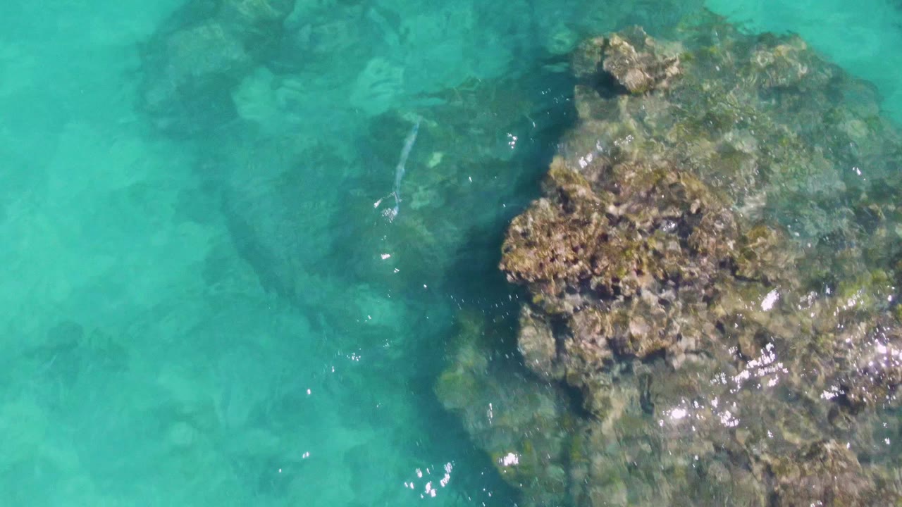 Overhead Buzzards Beach frame with a large submerged rock formation centered low in the image, pale turquoise water around it, and no visible people.
