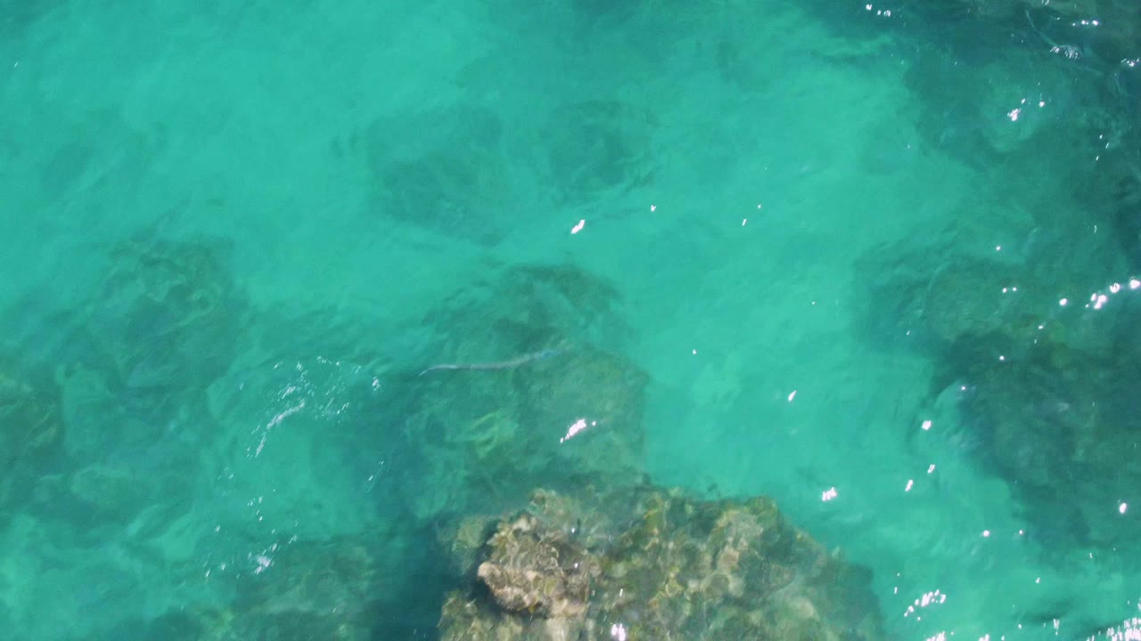 Overhead Buzzards Beach view with darker submerged rock shapes under turquoise water, small sun reflections on the surface, and no visible people.
