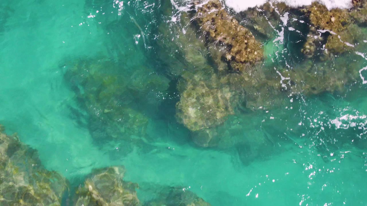 Top-down view of clear turquoise water over submerged rocks at Buzzards Beach, Baja California Sur, with small foam streaks and no visible people.