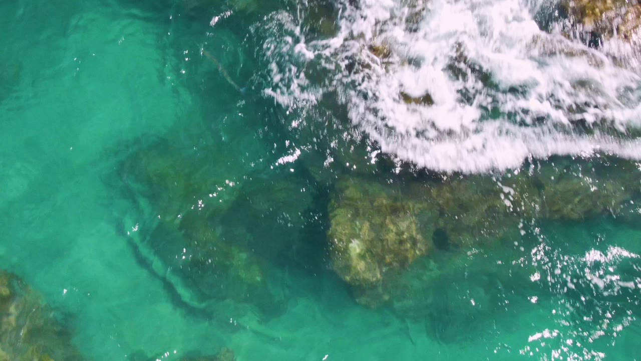 White wave foam enters the upper edge over brown-green submerged rocks in clear turquoise water at Buzzards Beach, with no visible people.