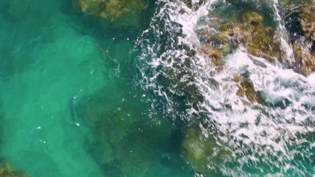 A larger strip of breaking white foam crosses the upper right over visible submerged rocks and green-blue water at Buzzards Beach, with no visible people.