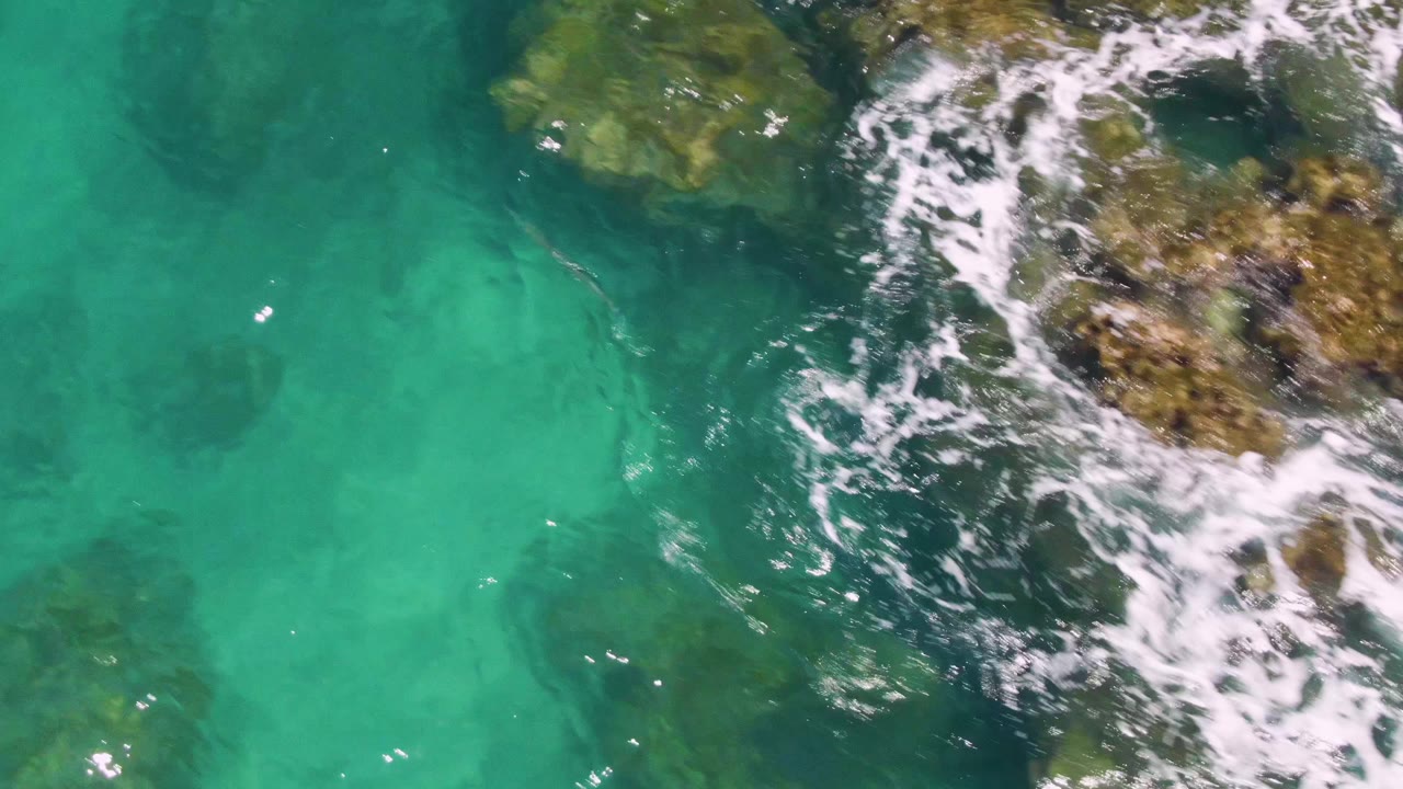 Clear turquoise water reveals darker underwater rock shapes while white foam washes along the right side at Buzzards Beach, with no visible people.