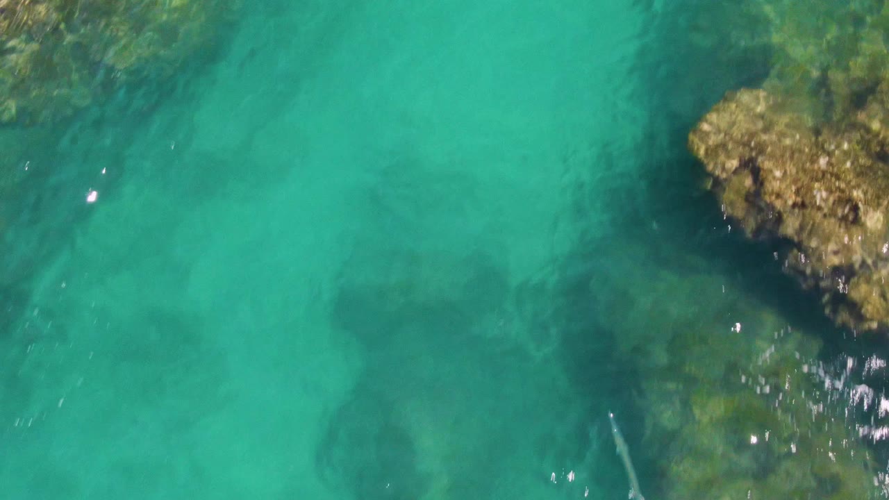 Overhead frame from Buzzards Beach showing a tan rock outcrop at the top edge, clear shallow water below it, and no visible people.