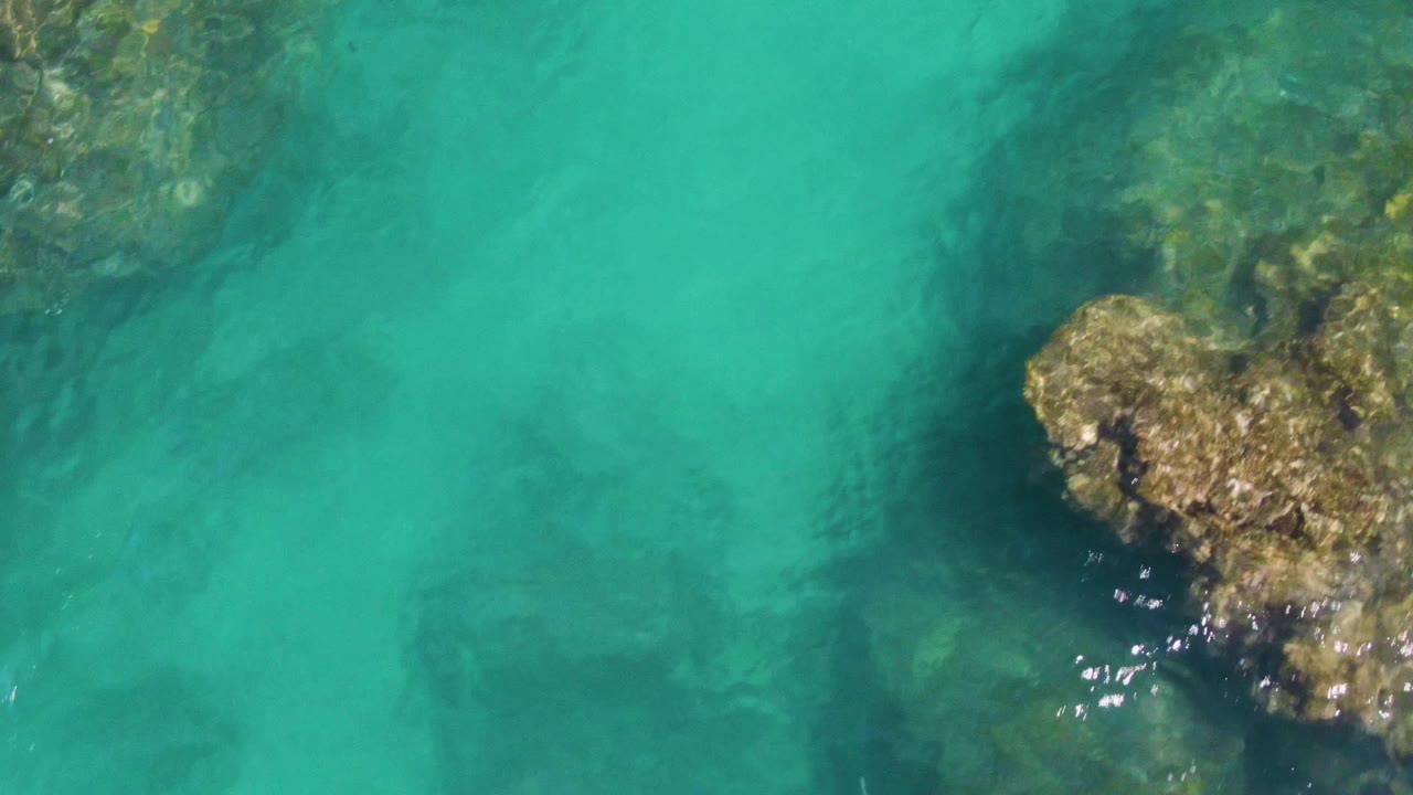 Overhead view at Buzzards Beach with exposed tan rock in the upper right, submerged rocky shapes under the turquoise water, and no visible people.