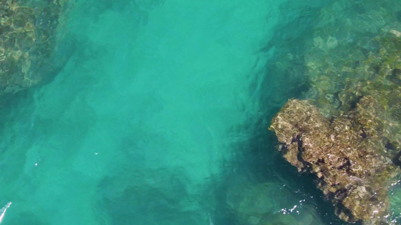 Overhead Buzzards Beach frame with a larger rocky formation crossing the center and lower right, clear turquoise water around it, and no visible people.