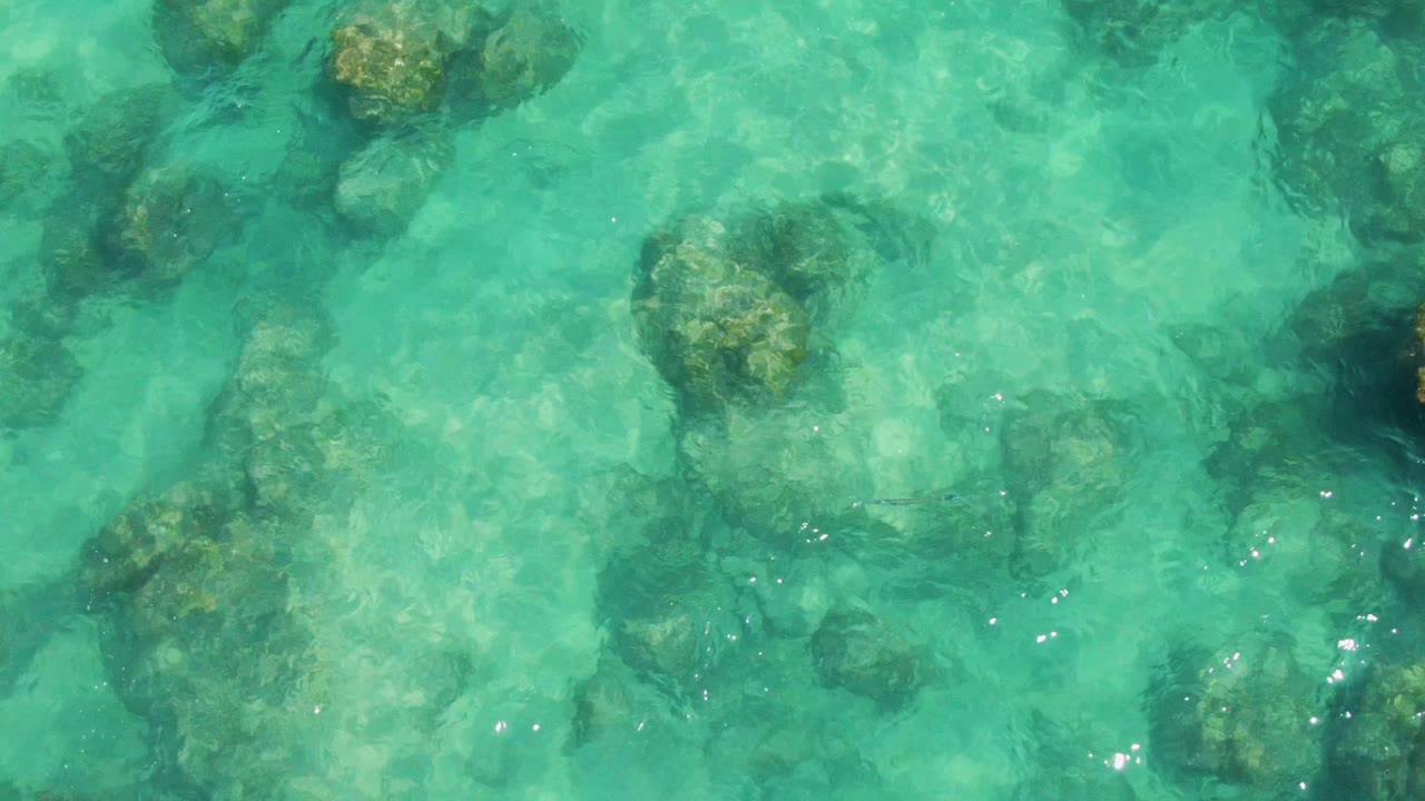 Clear green-blue ocean water at Buzzards Beach shows several rounded rocks beneath the surface, with soft ripples slightly bending the view and no visible people.