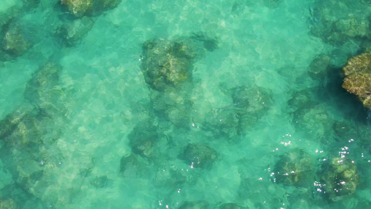 Clear turquoise sea at Buzzards Beach shows larger submerged rocks near the frame edges and smaller dark shapes across the seabed, with no visible people.