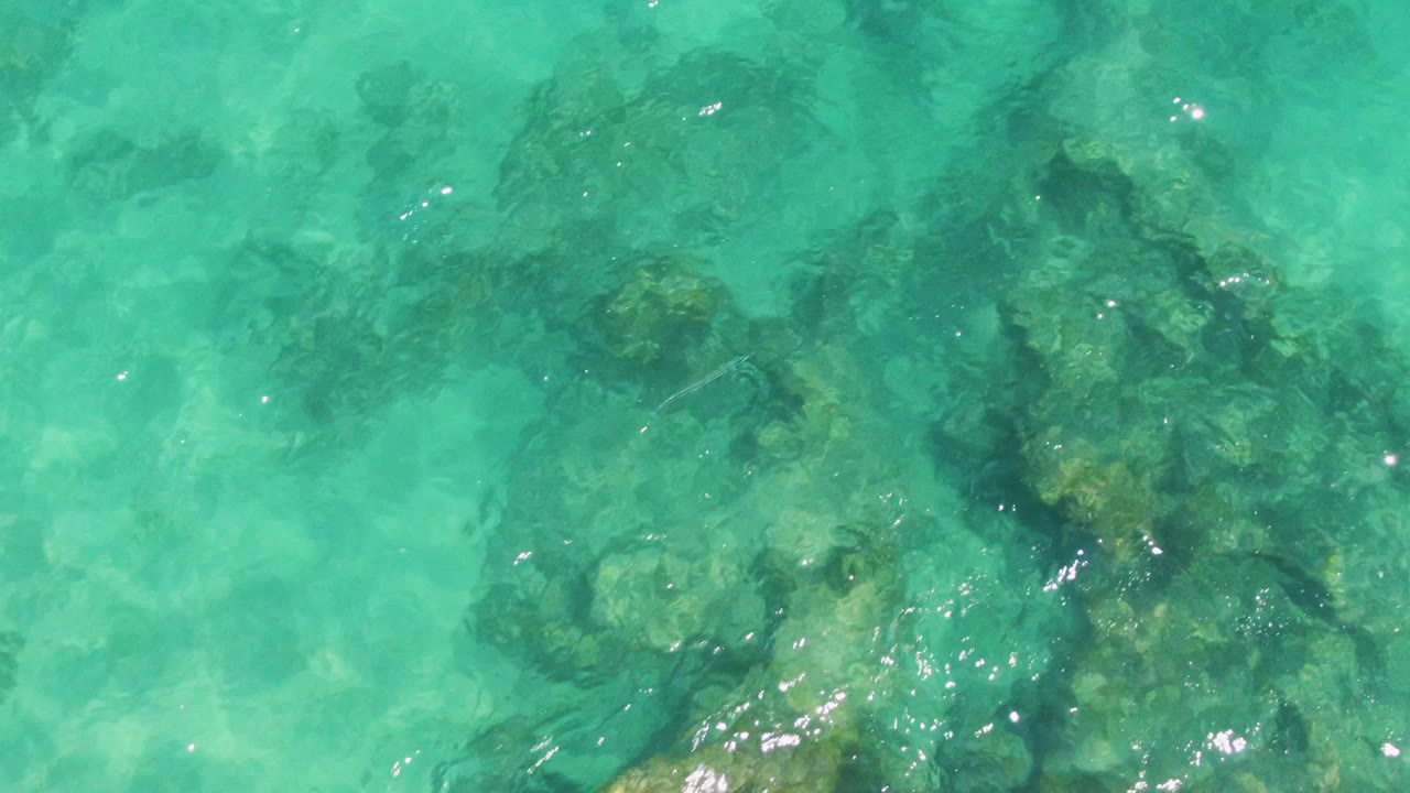 Clear green-blue water covers darker underwater rock patches at Buzzards Beach, Baja California Sur, with ripples softening the rock edges and no visible people.