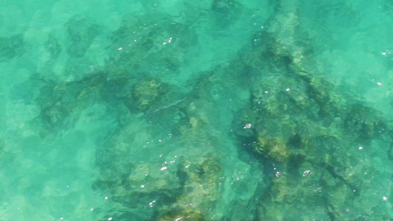 Shallow turquoise ocean at Buzzards Beach, Baja California Sur, reveals uneven rock formations under the surface, with bright highlights scattered across the water and no visible people.