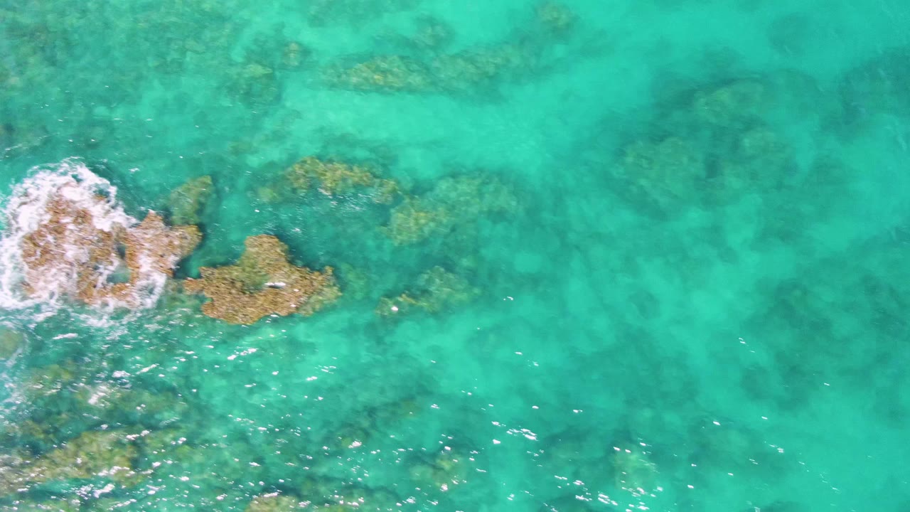 Aerial frame from Buzzards Beach showing white surf wrapped around a submerged rock cluster, with clear turquoise water and no visible people.