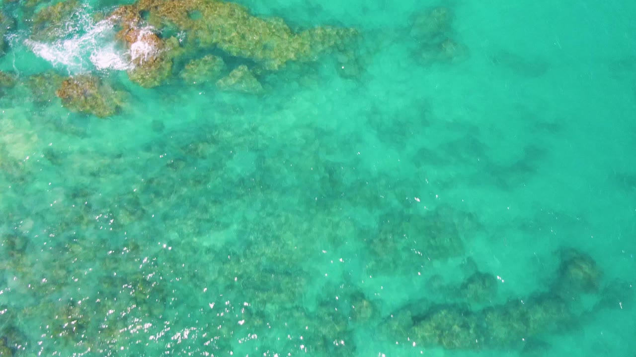 Overhead aerial frame from Buzzards Beach with a foamy rock patch along the top edge, darker submerged formations below the surface, and no visible people.