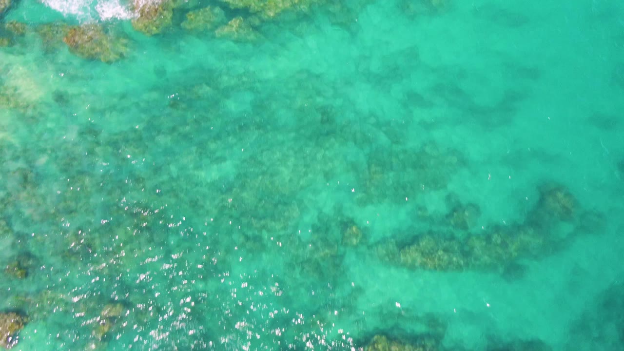 Top-down view at Buzzards Beach, Baja California Sur, with pale turquoise shallows, scattered submerged rocks, a small foamy area near the top left, and no visible people.