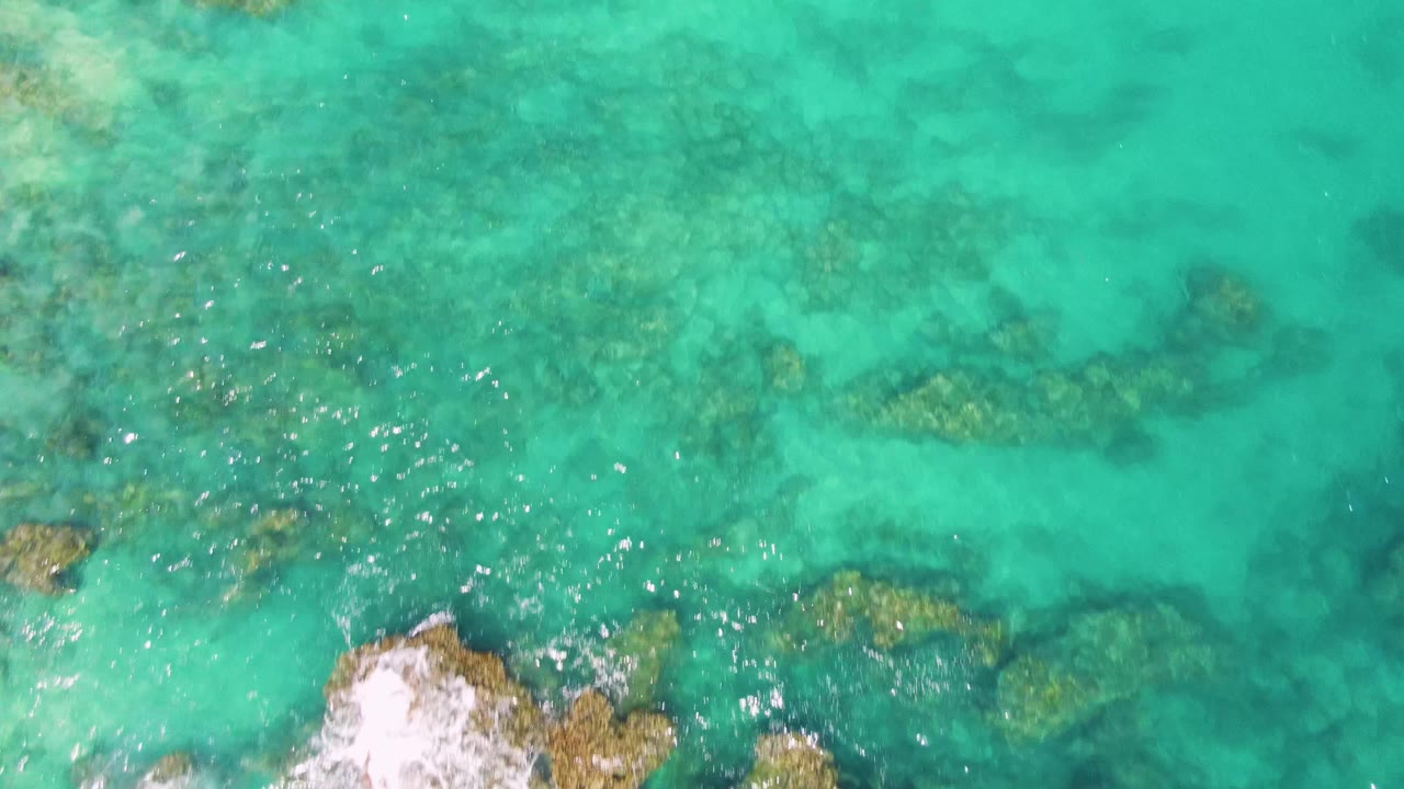Overhead frame from Buzzards Beach showing submerged rock bands across clear turquoise water, bright reflections on the left, white foam entering from the lower left, and no visible people.