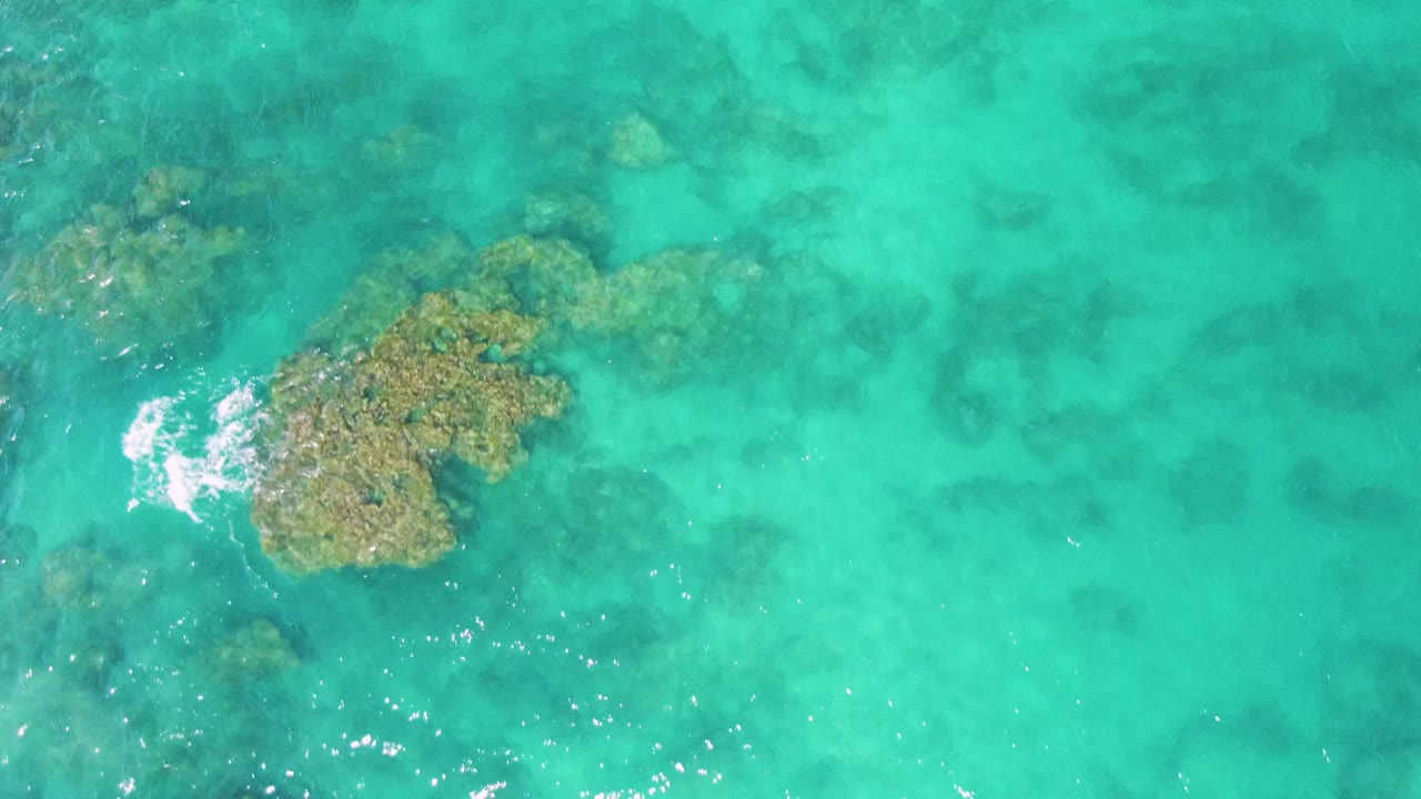 Overhead frame of turquoise water at Buzzards Beach with a large submerged rock cluster near the upper left, smaller rocks below, sun glints, and no visible people.