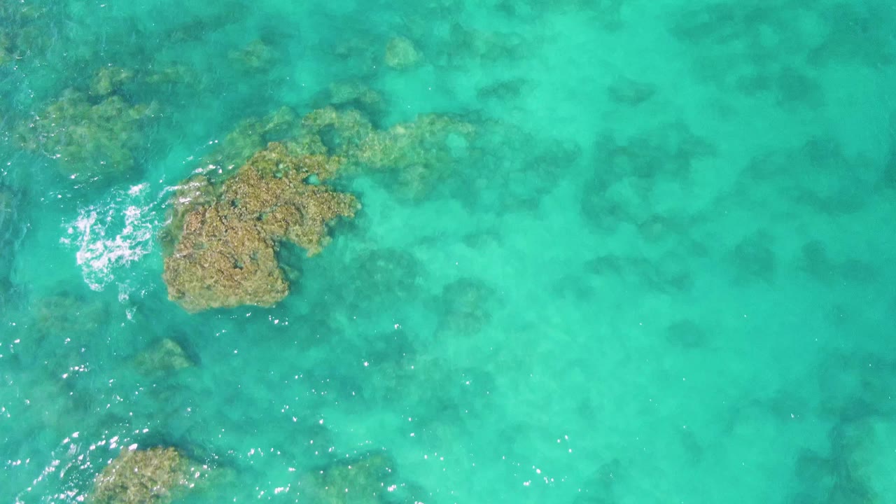 Overhead frame at Buzzards Beach showing a central rock formation under clear turquoise water, darker seabed patches, small white ripples, and no visible people.