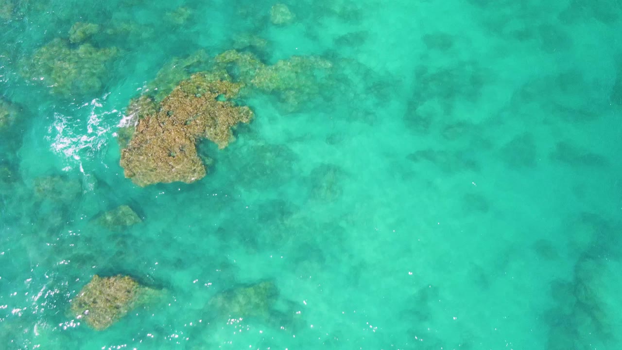 Overhead Buzzards Beach frame with a rock cluster near the upper right, smaller formations below it, bright turquoise water, scattered foam, and no visible people.