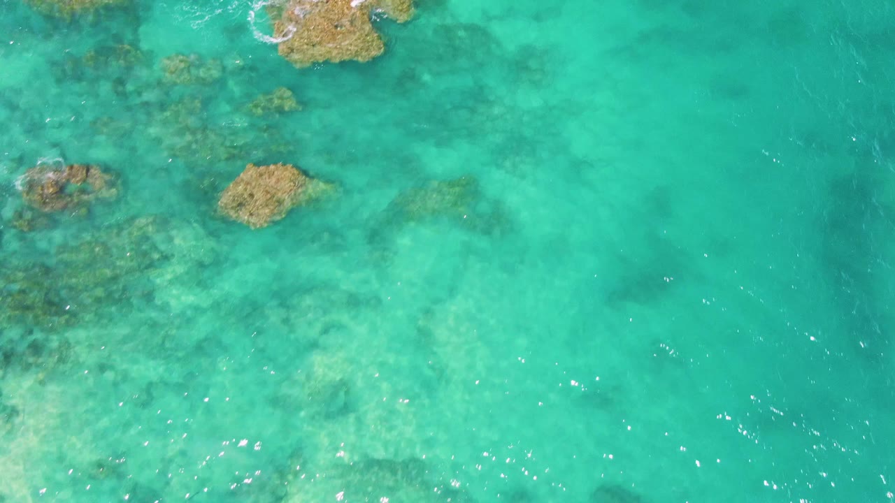 Overhead view of clear turquoise water at Buzzards Beach, Baja California Sur, with submerged rocks near the upper edge and darker reef patches below, no visible people.
