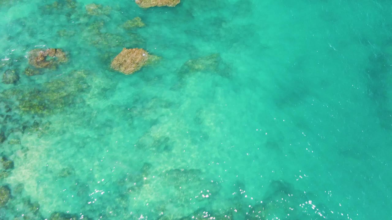 Clear green-blue water at Buzzards Beach reveals pale sand, darker submerged reef patches, and small surface highlights, no visible people.