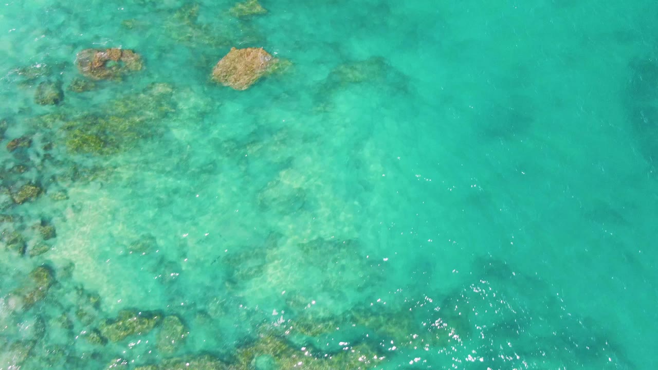 Overhead frame at Buzzards Beach showing turquoise shallow water, tan rock clusters, darker underwater patches, and bright sun flecks on the surface, no visible people.
