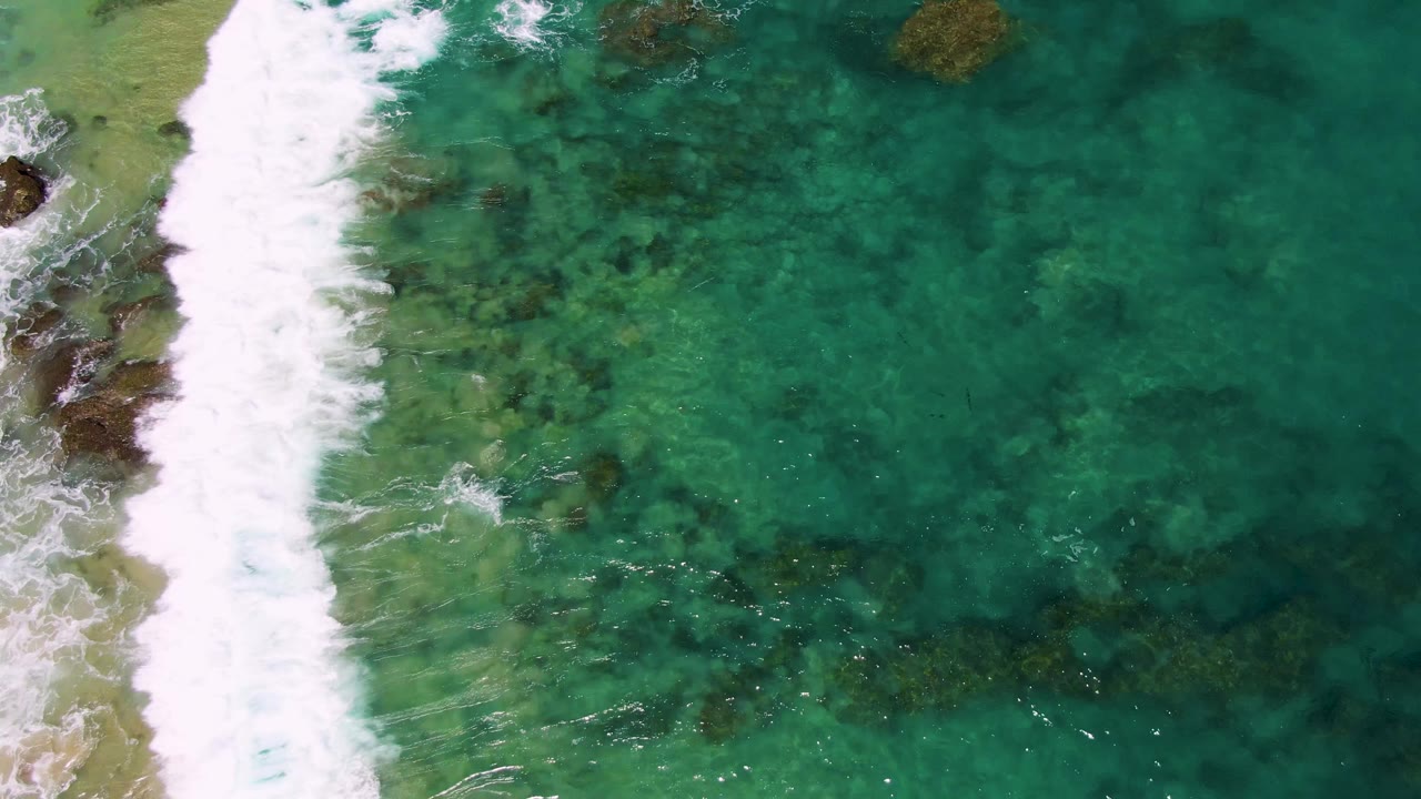 Overhead view at Buzzards Beach, Baja California Sur, with a thick band of white surf along the left edge and clear turquoise water revealing submerged rocks, no visible people.