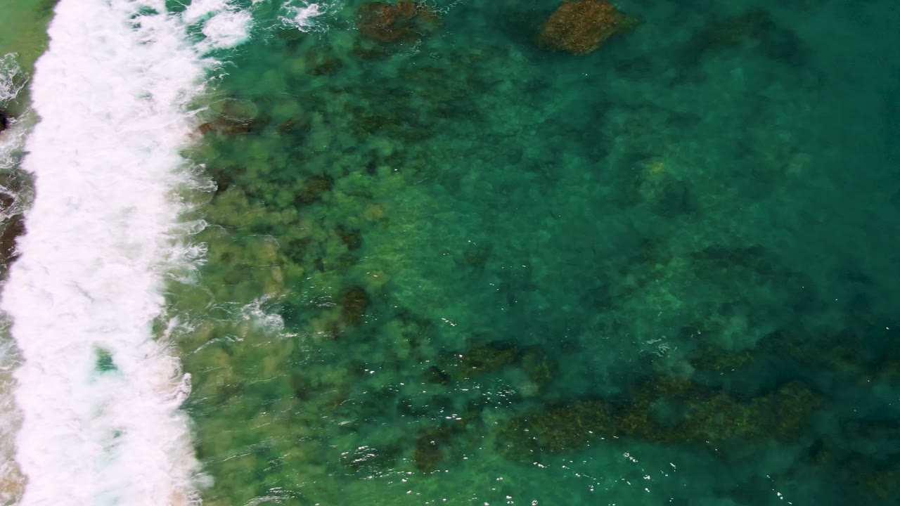 Foamy wave band crosses shallow green-blue water with rocks below.