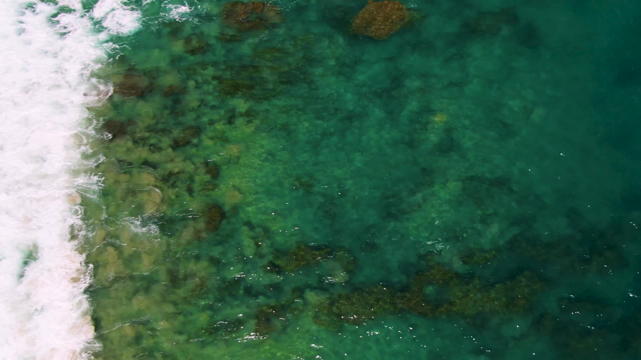 Straight-down drone view at Buzzards Beach with white surf along the left side, scattered brown rock patches beneath turquoise water, and darker open water to the right, no visible people.