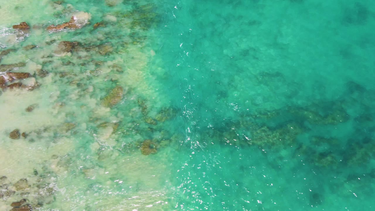 Overhead shoreline frame at Buzzards Beach, Baja California Sur, where pale sand and brown rocks sit under clear turquoise water with no visible people.