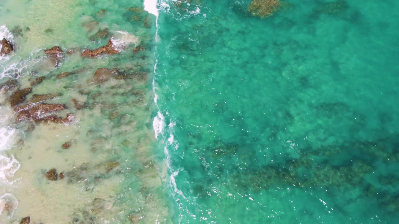 A narrow white wave line runs down the left side of the overhead frame beside rocks and pale sand at Buzzards Beach, Baja California Sur, with no visible people.