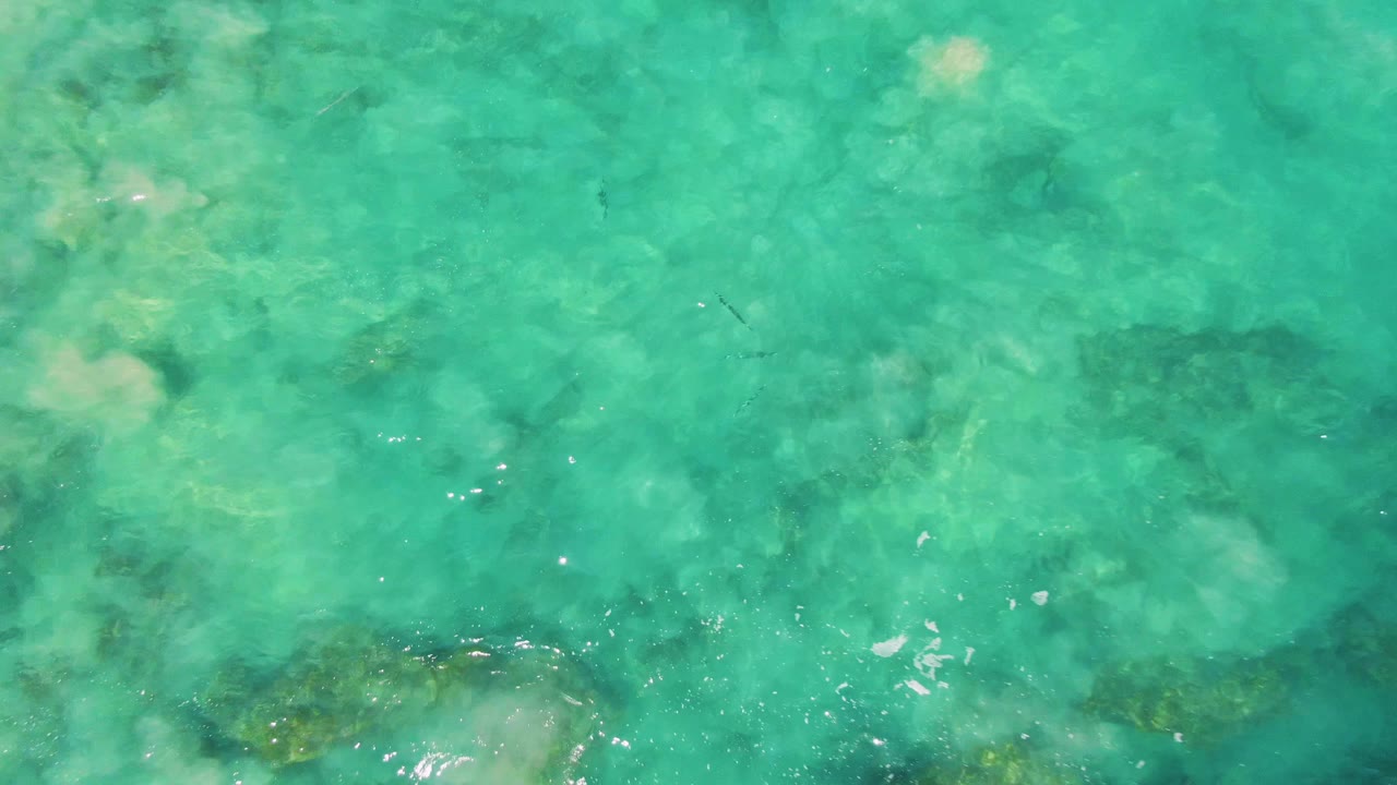 Clear shallow water at Buzzards Beach shows pale seabed zones, darker submerged formations, small ripples, and no visible people.