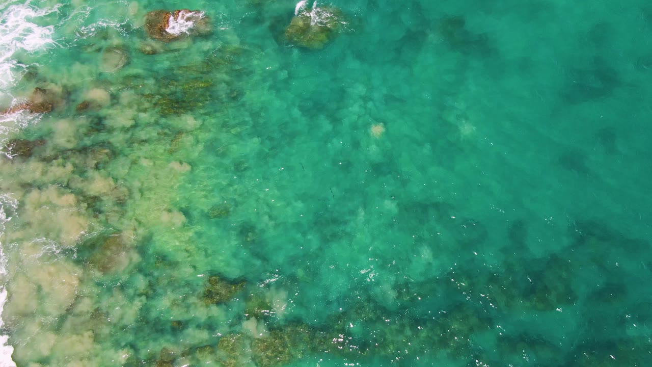 Overhead view of shallow turquoise water at Buzzards Beach, Baja California Sur, with submerged rocks near the top and left edges, pale sand below the surface, small white surf, and no visible people.