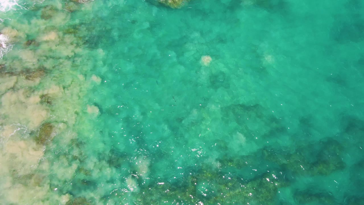 Overhead Buzzards Beach frame with turquoise water filling the view, pale sand and darker reef-like rock shapes below the surface, a few bright highlights, and no visible people.